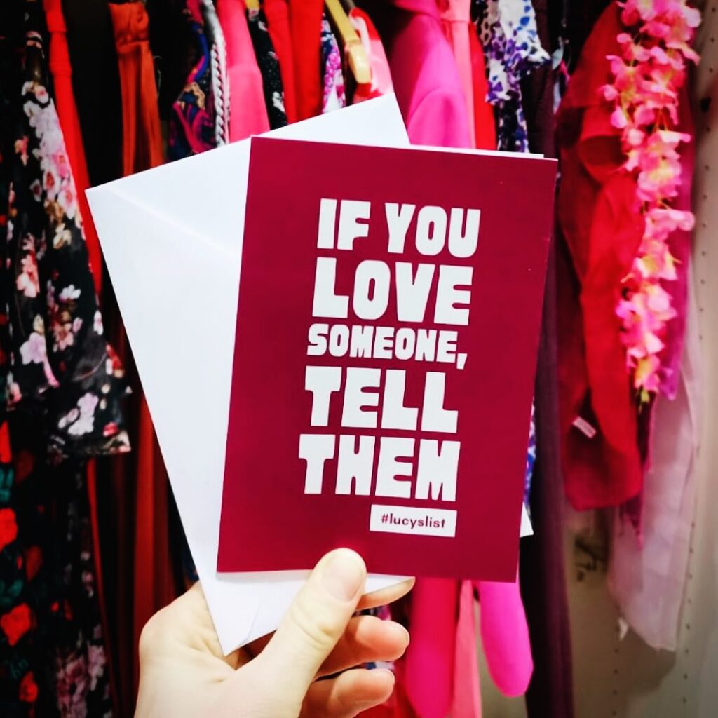 A card that reads "If you love someone, tell them #lucyslist" being held up against a backdrop of vintage clothes in an ethical boutique.